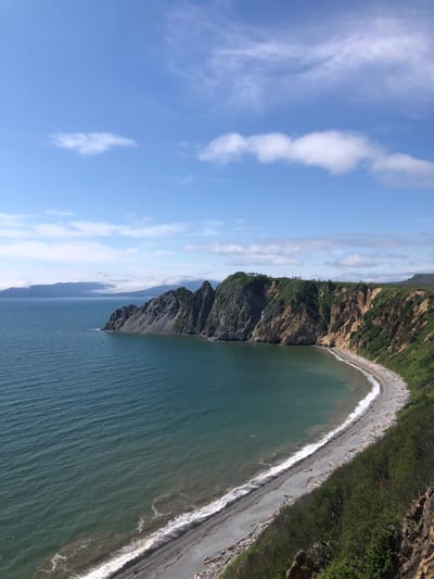 This is a high-angle, full shot of a rocky coastline on a clear day. The ocean is a deep teal with small waves lapping against a grey pebble beach. The beach is littered with driftwood. The cliffs are covered in green vegetation and some exposed rock faces. In the distance, hazy mountains are visible. The sky is bright blue with scattered white clouds. The scene is located in the city of Ola, Russia.