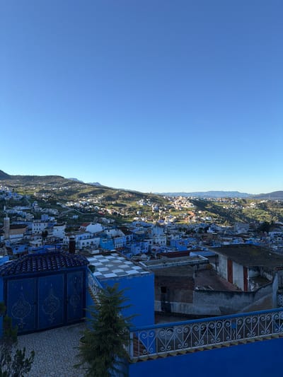 An elevated, wide-angle image captures a daytime panorama of Chefchaouen, Morocco, under a clear, cloudless blue sky. In the foreground, a blue tiled terrace is visible, featuring a dark blue, ornate structure with two arched, patterned doors and a ridged, dark blue tiled roof on the left. Adjacent to it on the right is a blue building with a flat roof displaying a white and blue checkerboard pattern. A small, green evergreen plant is positioned between these structures, with a white, decorative iron railing on a blue base visible on the far right. The midground shows the dense urban landscape of Chefchaouen, characterized by buildings predominantly painted in various shades of blue and white, with some structures showing exposed brick or concrete. A slender, light-colored minaret with a pointed cap rises from the cityscape on the left. The background features extensive green, undulating hills dotted with scattered white and light-colored buildings, extending to distant mountains under bright sunlight. No individuals or active scenes are depicted.