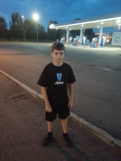 A young boy stands in the foreground of a gas station at dusk, likely in Alessandria, Italy. He is wearing a black t-shirt with a blue cartoon owl graphic and the text "SAMBOYS" underneath, and black shorts. He is also wearing black socks and shoes. The boy is facing the camera with a neutral expression.
Behind the boy, the gas station is illuminated by overhead lights. Several gas pumps are visible, along with the canopy of the station. The sky is a deep blue, indicating it is either dawn or dusk. Trees line the background of the scene, and a street lamp is visible to the left. The ground is asphalt, with a curb and a metal grate on the left side of the image. The overall atmosphere is calm and quiet, consistent with the late hour. There are no other people or vehicles immediately visible.