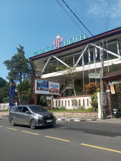 The image shows the Palace Hotel in an urban setting, likely in Indonesia based on the car license plate. The hotel has a sign with "PALACE HOTEL" in green lettering and a logo with a crown above the letters "PR". A gray minivan with license plate "B 1431 CZL" drives on the asphalt road with yellow lines in front of the hotel. There are trees and greenery surrounding the hotel. A sign for Bank Artha Grama Internasional (AGI) stands near the entrance. Another sign advertises "PUSAT OLEH-OLEH LEGENDARIS PUNCAK" with an image of Lobby Lounge Rambyn Cafe. A smaller sign reads "BINTANG TERANG". The sky is blue with some clouds.