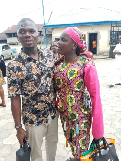 Two people are standing in front of a building in Yenagoa, Nigeria. The man is wearing a floral shirt, khaki pants, and a black leather bag over his left shoulder. The woman is wearing a pink and yellow patterned dress with a pink long-sleeved shirt over it and a pink head wrap. The man is holding the woman's right arm, and she is looking to the right while the man is looking at her. They are standing on a brick pathway, and there is a blue and white building behind them. Another woman is standing behind the man, looking to the right. A car is parked on the left side of the image. The man is holding a black bag in his left hand. The woman has a black purse on her left shoulder and is holding a bag in her right hand. The building is a light blue and white building, with a blue roof. There is a gate in the front of the building with a light blue door behind it. The scene is taken outside. The weather is sunny. The image was likely taken during the day.