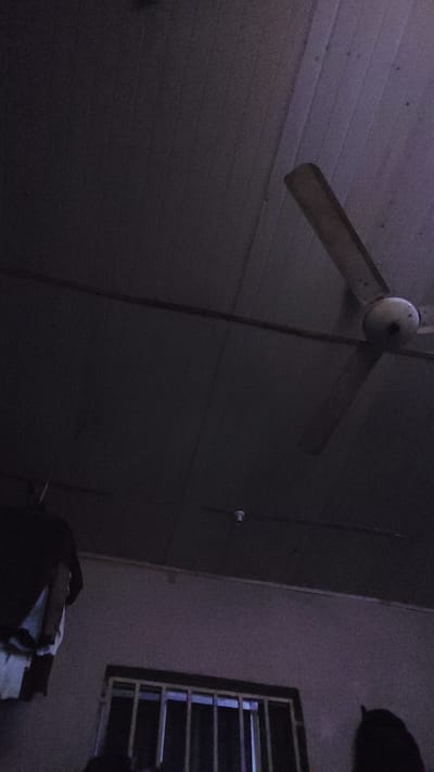 The image depicts an interior scene, likely a room, with a ceiling fan visible. The lighting is dim, suggesting it might be evening or a poorly lit space. The fan is mounted on the ceiling, and its blades are spread out. To the left of the image, there is a window with bars, a common security feature. The walls appear to be paneled or textured. A dark object, possibly furniture or a person's silhouette, is present in the lower left corner. The overall atmosphere is subdued due to the low light conditions. No specific activity or event is clearly identifiable. The location context of New Nyanya, Nigeria, provides a geographical reference. There is no visible text in the image.