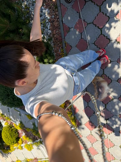 A low-angle, full-body shot captures a person from the perspective of someone holding a camera overhead. The person is wearing light blue jeans and a grey t-shirt, with red flip-flops on their feet. They are walking on a paved path with a decorative pattern of grey and pink tiles. In the background, a small, fluffy dog on a leash is visible, appearing as a blur. The scene is outdoors, with the sunlight casting shadows on the ground, indicating it is daytime. The setting appears to be a garden or yard, with various plants and greenery surrounding the path. A metal chain, likely attached to the dog's leash, runs diagonally across the frame. The person's arm is also visible, adorned with a silver bracelet. The overall impression is of a casual outdoor stroll.