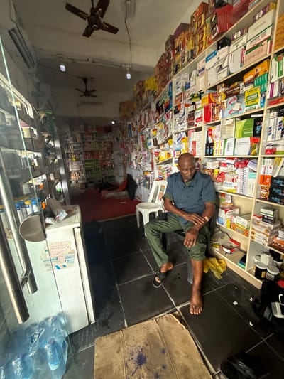 The image shows a man lying on the floor of a pharmacy, seemingly asleep or resting. He is wearing a blue shirt and green trousers. The pharmacy shelves are filled with various medications, boxes, and bottles. Some visible text includes brand names like "coskin," "Erythrofaith," "ANACIN," "BERLIN FLUCONAZOLE," and "MAN-G." The setting appears to be indoors, likely a small retail space, with fluorescent lights and ceiling fans visible in the background. The floor is tiled in black. The overall impression is of a busy but perhaps somewhat cluttered pharmacy, with the man in a relaxed, reclined posture amidst the merchandise. The image is oriented sideways, suggesting it was captured with a mobile device.