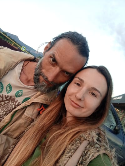 A couple is captured in a selfie against an outdoor, possibly rural, backdrop. The man, in the foreground and to the left, has a beard and dark hair tied back. He wears a light-colored t-shirt with a green leaf pattern and a tan jacket. His left eye is visible and he is looking directly at the camera. The woman is to the right, leaning her head towards the man. She has long, light brown hair and appears to be smiling faintly. She is wearing a green garment and a fur-trimmed vest. In the background, mountains are visible under a cloudy sky. A yellow road sign with a black 'A' is present on the left side of the image, suggesting a roadway or public area. A portion of a dark-colored car is visible on the right.