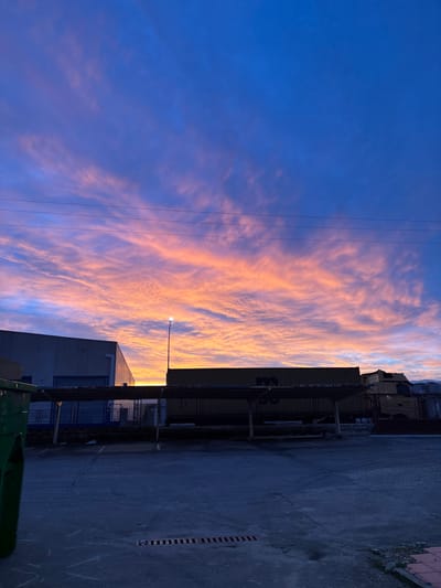 An exterior image depicts an industrial or commercial area in Valverde del Majano, Spain, at twilight. The sky transitions from deep blue overhead to vibrant orange and pink altocumulus clouds near the horizon. The foreground consists of a dark, empty asphalt or concrete parking lot with a rectangular metal drain grating present. A green commercial dumpster is partially visible on the far left. In the midground, a light-grey industrial building with a flat roof and a large recessed opening protected by blue grating is situated on the left. Centrally, a tall pole with an illuminated street lamp fixture stands. To the right of the lamp, a dark, low-lying canopy or covered structure extends horizontally, beneath which a yellow-tan cargo container with dark markings and parts of other industrial vehicles, including yellow-tan truck cabs, are visible. Multiple dark utility lines traverse the upper portion of the sky. No individuals are present in the scene.