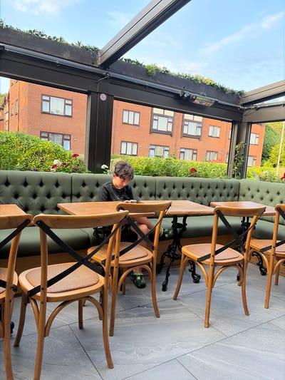 An exterior shot of a covered outdoor seating area with a young male seated at a table. The seating area features a long, tufted, dark green upholstered bench and wooden chairs with woven seats. Several small wooden tables with dark metal bases are present. The young male, wearing a black t-shirt and green sneakers, is looking down at a mobile phone in his hands. Behind the bench, a dense hedge with scattered red and white flowers is visible, and beyond that, a red brick building with multiple windows. The sky is visible above the covered structure, which has a dark frame and a clear roof. The overall scene suggests an outdoor dining or cafe setting. This image was taken in Salford, United Kingdom.