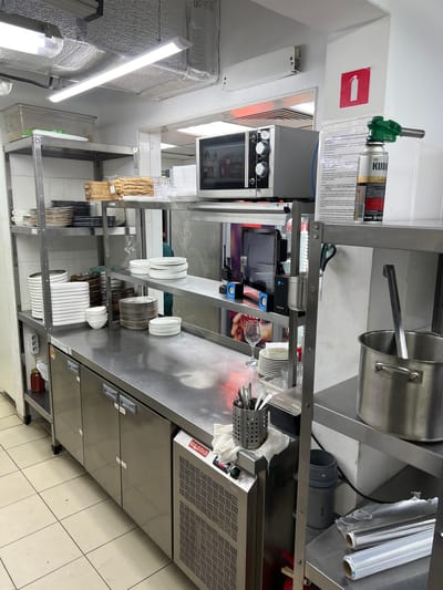 The image depicts a commercial kitchen environment, specifically a prep or service area, in Plastunivska, Russia. Stainless steel shelving units dominate the scene, laden with a variety of kitchenware including stacks of plates, bowls, and cutlery. A microwave oven is visible on a shelf, alongside a gas torch with a can labeled "KUDO." Refrigerated units are present beneath a stainless steel countertop, some with doors displaying labels. The floor is tiled with light-colored squares. A fire extinguisher is mounted on the wall, and other items like jars of sauce, rolls of plastic wrap, and a large metal pot with a ladle are also present. The overall impression is of a functional, albeit somewhat cluttered, back-of-house operation. No individuals are clearly visible in the frame.