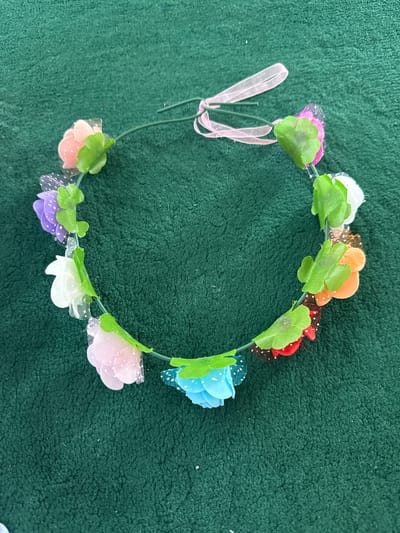 A flower crown made of small fabric flowers in various colors, including pink, purple, white, orange, red, and blue, is laid out on a dark green, textured surface. The crown has a green wire base and is adorned with green fabric leaves. A pink ribbon is tied to one end of the wire. The image is an overhead shot, with the flower crown forming a partial circle. The green surface appears to be a carpet or similar fabric. There are no people or buildings visible in the image. The location of the image is not determinable from the visual information provided.