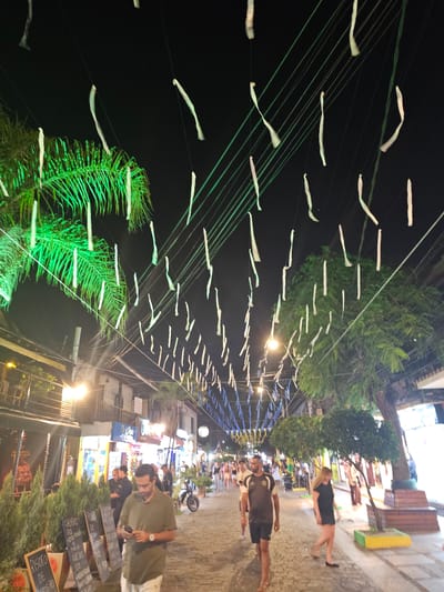 A bustling street scene at night in Armação Dos Búzios, Brazil. Paper decorations are strung across the street, illuminated from below by street lamps and business lights. People are walking along the sidewalk, some in groups, others alone. They are dressed in casual clothing, like shorts and shirts. Palm trees and other greenery are scattered throughout the scene, adding a touch of nature to the urban environment. Restaurant menus are displayed outside an establishment on the right side of the street, suggesting a vibrant dining scene.
