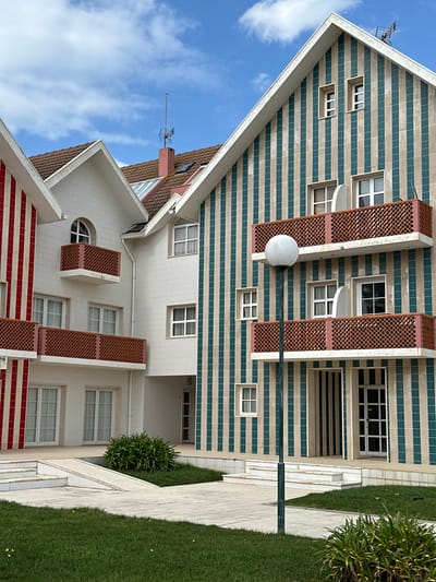 The image depicts a residential complex in Ílhavo, Portugal, characterized by multi-story buildings with prominent gabled roofs and distinctive striped facades under a partly cloudy blue sky. The central building is off-white, featuring multiple rectangular windows and one arched window, each with a small reddish-brown latticework balcony. To its left, a section of a building exhibits vertical red and white stripes with a similar balcony. To the right, a larger building displays vertical teal/green and beige stripes across its facade, featuring a variety of window sizes and two extended balconies with reddish-brown latticework railings. A white-framed glass door is visible at ground level on this striped building. The roofs are covered with brown tiles, and a reddish-brown chimney with an antenna and another smaller antenna are present on the rooftops. The ground level consists of green grass areas interspersed with concrete pathways, stairs, and a ramp. Green shrubbery is planted in designated sections of the landscaping. A dark-colored lamppost with a spherical white light fixture stands in the mid-ground. No individuals are visible.