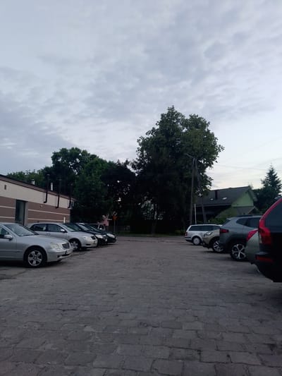 The image captures a cloudy sky over a parking lot paved with gray cobblestones. Several cars are parked in a row. On the left, a silver sedan is visible, followed by other sedans and SUVs. A building with a light brown facade and a dark roof is situated to the left of the parking lot. Behind the parking lot, lush green trees and a two-story green house with a dark roof are visible. A wooden utility pole stands among the trees, with power lines running across the sky. The scene appears to be a quiet residential or commercial area in Wołomin, Poland, during the late afternoon or early evening.