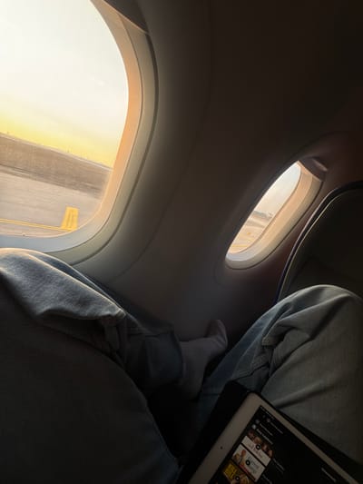 The image captures the interior of an aircraft at Ben Gurion Airport (TLV) in Yehud Monosson, Israel, observed from a passenger's seat. The lower body of a single individual is visible, wearing light blue denim jeans. Their left foot is covered by a white sock. A black digital tablet, identified as an iPad, rests on the person's lap, displaying a streaming service interface with movie thumbnails including "Kill Bill," "Forrest Gump," "The Holiday," and "Sexy Beast." Two oval-shaped aircraft windows are prominent, framed by the light grey interior paneling of the cabin. Through the windows, an airport tarmac is visible under a bright, golden sky, consistent with either sunrise or sunset. The tarmac shows asphalt pavement with yellow ground markings, and a distant cityscape is faintly discernible on the horizon.