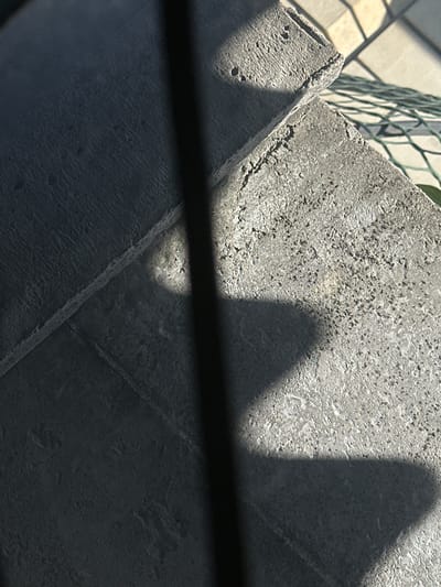 In Lobacheve, Ukraine, the image captures a close-up view of two rough, gray concrete paving stones set against a background of lighter, patterned paving and a hint of green mesh. The stones are arranged at an angle, with one partially overlapping the other. Sunlight casts sharp shadows across the scene, creating a play of light and dark that highlights the textured surfaces of the concrete. A dark, vertical shadow line bisects the image, suggesting an obstruction like a fence post or a thin structure. The overall impression is one of an outdoor, possibly urban or suburban, environment with a focus on the materiality of the concrete elements and the effect of natural light.
