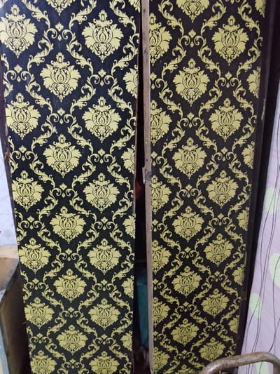 The image shows a close-up view of two cabinet doors with a black background and a repeating yellow damask pattern. The pattern consists of ornate floral motifs and swirling vines. The doors are slightly ajar, revealing a glimpse of what might be inside. The cabinet appears to be a piece of furniture, possibly a wardrobe or cupboard, standing in an indoor setting. The lighting suggests it could be daytime, but there are no strong indicators of the time of day. The overall impression is one of a decorative interior element. There is no visible text in the image.