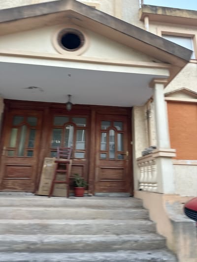 The image shows the facade of a building in the city of Seoce, Montenegro. The building appears to be a multi-story structure with a light-colored stucco exterior. In the foreground, there is a set of concrete stairs leading up to a covered porch. The porch has a white ceiling and is supported by columns. Two large, dark wooden doors are visible on the porch, each with glass panes. To the left of the doors, a red chair and a cardboard box are placed on the steps. A potted plant sits to the right of the doors. The roofline above the porch is triangular and features a circular, dark opening. To the right of the entrance, a window covered with a roller blind is visible. The overall scene suggests a residential building, possibly undergoing renovation or with items stored outside.