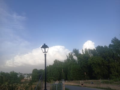 The image depicts an outdoor scene in Navaluenga, Spain, featuring a streetlamp in the foreground, a line of lush green trees in the middle ground, and a cloudy sky with large cumulus clouds in the background. A person is walking on a paved path to the right of the frame, accompanied by a dog. The environment suggests a rural or semi-rural setting, with vegetation and a residential building visible behind a fence in the distance. The lighting indicates it is daytime, likely late afternoon or early evening given the sky conditions.