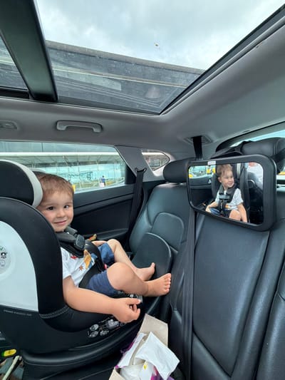 The image shows a young child secured in a car seat inside a vehicle. The child is wearing a white t-shirt and blue shorts. The car has a sunroof that is open, revealing a cloudy sky. In the background, visible through the car windows, is an airport terminal building with people and luggage. The specific location is identified as El Prat de Llobregat, Spain, which is the municipality where Barcelona Airport is situated. A mirror is attached to the headrest of the front seat, reflecting the child in the car seat.