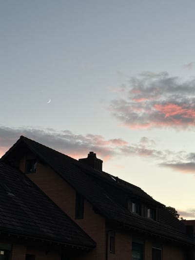 This is a photograph taken from a low angle, looking up at the sky and the roof of a building in Lufingen, Switzerland. The sky is a gradient of pale blue at the top, fading to a lighter, almost yellowish hue near the horizon. There are clouds in the sky, some appearing dark and heavy, while others are tinged with pink and orange from the setting sun. A crescent moon is visible in the upper left quadrant of the image.
Dominating the lower portion of the frame is the dark silhouette of a pitched roof, covered in dark tiles. The roof belongs to a brick building, the reddish-brown brickwork visible on the upper sections. The building has several windows, some appearing dark and reflective, others with shutters. A chimney is visible on the peak of the roof. The overall lighting suggests it is dusk or early evening. No people are visible in the image.