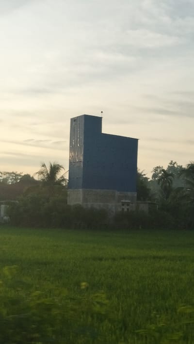 A tall, blue, rectangular building with a shorter, wide section attached to its right stands against a hazy, overcast sky. The building's base appears to be constructed of concrete blocks, with some visible windows. Lush green foliage and trees surround the base of the structure, with a dense line of bushes in front. A vibrant green field, likely rice paddies, occupies the foreground, stretching across the bottom of the image. The overall scene suggests a rural or semi-rural environment in Meunasah Ara, Indonesia.