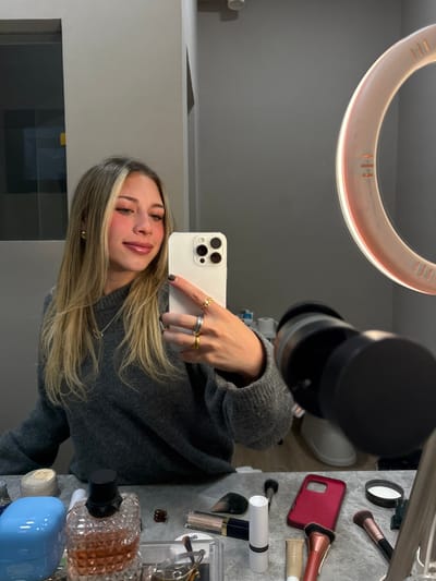 An image captured via a mirror reflection depicts a young female taking a selfie indoors. The specific location within Monterrey, Mexico, cannot be determined from the visual information provided.
**Subject and Action:** A light-skinned female with long, blonde-highlighted hair, wearing a gray knit sweater, gold stud earrings, a silver necklace, and multiple rings on her right hand, is holding a light-colored smartphone (equipped with a triple-lens camera array) to capture her own reflection. She is smiling subtly with light makeup applied to her face, including blush and lip gloss.
**Environment and Objects:** The female stands before a mirror reflecting an indoor setting, likely a room with light gray walls. A vanity surface or tabletop in the foreground is cluttered with various cosmetic items. These include multiple makeup brushes, a red smartphone case, a large faceted perfume bottle (appearing to be Valentino), a blue container, several tubes and compacts of makeup (e.g., lipstick, powder, foundation), and a small brown hair clip. To the right, a large circular LED ring light, mounted on a stand, is visible, emitting a glow that illuminates the scene. The reflection of a white, possibly ceramic, object is also visible in the background, behind the ring light.
