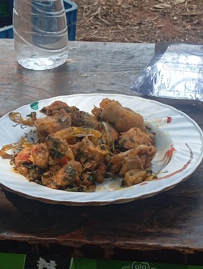 A close-up photograph depicts a meal set outdoors in Egede, Nigeria. The primary subject is a white, fluted ceramic plate holding a portion of stewed food, which appears to consist of meat or poultry pieces, green leafy vegetables, and sliced red and yellow peppers. To the left of the plate, a clear plastic bottle containing approximately 50% water stands on a weathered wooden table surface. To the right, partially visible behind the plate, is a silver-colored package with "NO. A1-106526L" printed on it. The background consists of dry, reddish earth sparsely covered with dry leaves and debris. A blue plastic container is partially visible in the upper left background. The extreme foreground shows a green surface with a partial 'glo' logo. No individuals are present or engaged in any direct action or interaction within the frame.
