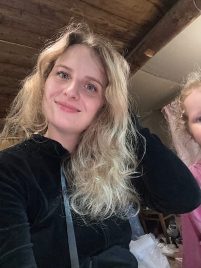 A medium close-up selfie shows a fair-skinned woman with blonde, wavy hair. She is wearing a black hooded jacket. To her right, a child with blonde curly hair is partially visible, wearing a pink garment. The background appears to be an indoor setting with a wooden ceiling and a cluttered floor with bags and shoes. The location is not specified in the image or metadata, and cannot be determined.