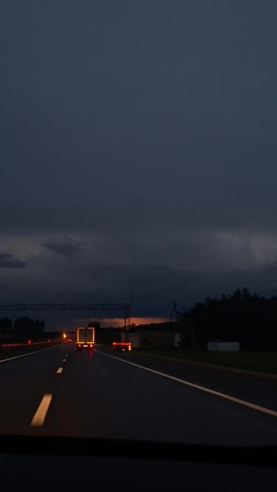 The image shows a nighttime scene on a highway in Самахвалавіцкі сельскі Савет, Belarus. The sky is dark and cloudy, with a hint of orange on the horizon. A vehicle with lit orange rectangular frame is moving along the road, following by the red lights. Road markings, including white lines and reflectors, are visible. The surrounding environment includes trees and potentially buildings in the distance. The asphalt road is smooth.