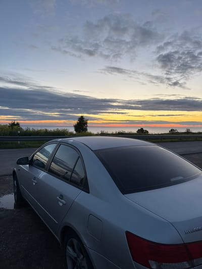 A silver sedan is parked on a gravel roadside facing a body of water at sunset. The sky is a gradient of blue, gray, and orange, with scattered clouds. The water appears calm, with a distant tree line. A guardrail runs along the edge of the road. The car is clean and appears to be in good condition. The scene is peaceful and serene. The location is identified as Paldiski, Estonia.