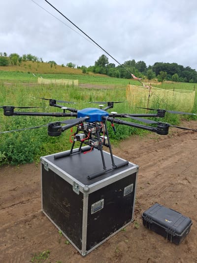 A large, blue and black drone with eight rotors is positioned on top of a large black flight case. The drone has four arms extending outwards, each with two rotors. The flight case has silver metal accents and latches, with the word "MULTIROTOR" visible on its side. To the right of the flight case, a smaller black case is lying on the ground. The scene is set in an outdoor environment with dirt ground and green vegetation. In the background, there is a field with crops, trees, and a yellow safety fence. A wire stretches across the top of the image. The sky is overcast and cloudy. The location is identified as Lubnica, Poland. There are no people visible in the image.