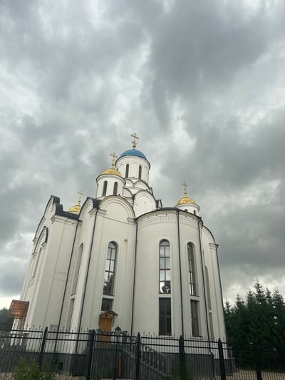 The image shows a white church with golden domes and crosses against a backdrop of a cloudy sky. The church has a traditional architectural style with arched windows and intricate decorative elements. A black metal fence runs along the side of the building, and a few trees are visible in the background. The sky is overcast, suggesting a cloudy or possibly rainy day. The lighting is diffused, indicating it is likely daytime, though the lack of direct sunlight creates a somewhat somber mood. The perspective is from a low angle, looking up at the church. No people are visible in the image. The scene is serene and solemn, characteristic of a religious building.