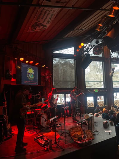 A live band performs in a dimly lit, rustic venue with exposed wooden beams and large windows. The band includes a guitarist, drummer, and possibly other musicians. Stage lighting casts a red hue. An audience is visible in the background. A vintage advertisement is visible on a wall. A screen displays information about Aaron Davis and a QR code for tips.