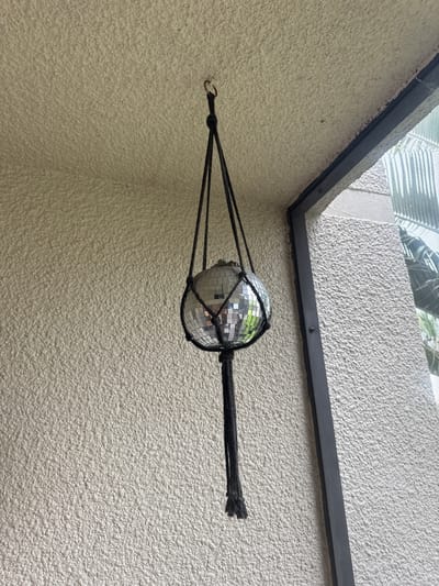 A disco ball, suspended from a black macrame hanger, hangs in front of a textured, light-colored wall. The disco ball is composed of numerous small, reflective square mirrors. The hanger is attached to a ceiling hook. To the right of the disco ball, a dark-colored window frame is visible, with a screen and the reflection of greenery beyond it. The setting appears to be an outdoor or semi-outdoor space, possibly a balcony or porch, given the wall texture and the presence of a window and screen. No people or animals are present in the image. The scene is static and shows a decorative object.