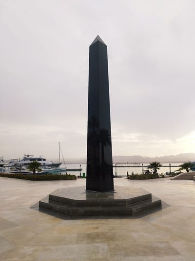 The image shows a tall, black, obelisk-like monument in Ras Abu Soma, Egypt. The structure has a square base that tapers to a point at the top. It appears to be made of polished stone or metal. The monument stands on a multi-tiered platform made of a similar dark material, set on a plaza paved with light-colored stone.
In the background is a harbor with several boats docked. Palm trees are planted along the waterfront. The sky is overcast and the light is soft, suggesting either early morning or late afternoon. The backdrop consists of distant mountains. The scene is devoid of people.