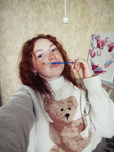 An overhead shot captures a young woman with reddish-brown, wavy hair, fair skin, freckles, and light blue eyes, looking upwards towards the camera. She is wearing a light-colored, possibly white or light grey, turtleneck sweater featuring a prominent brown and beige teddy bear design.
She holds a blue-handled paintbrush between her lips and chin, appearing pensive or thoughtful. To her lower right, an art canvas or board displays a floral pattern, possibly tulips, rendered in shades of pink, red, purple, and blue. The background is a textured, light-colored surface, suggesting an indoor setting like a wall or carpet. In the upper right corner, a white rectangular object, possibly a light switch or electrical outlet, is visible with a white wire extending from it. No text is visible in the image. The scene conveys a moment of quiet artistic contemplation in a domestic environment.