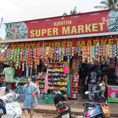 The image shows the exterior of Kavita Super Market in Oxel, India. The market's red awning displays its name in yellow, along with religious inscriptions. The entrance is lined with hanging packets of snacks and chips. Customers are visible, including a woman in a blue dress carrying a child. Several scooters are parked in front of the market.
