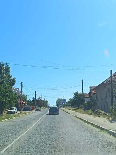 The image captures a sunlit scene on a two-lane asphalt road in Borovan, Bulgaria, likely during the daytime. The road is paved with textured asphalt and divided by a white line. The sky is a clear, bright blue, suggesting fair weather.
On the left side of the road, a sidewalk runs alongside residential buildings and mature trees with dense green foliage. Several parked cars are visible: a white hatchback and a dark sedan. Further down the road, utility poles with a complex network of wires stretch across the scene.
The right side of the road features a sidewalk adjacent to a building with a tiled roof and a light-colored facade, showing visible windows. A pile of bricks or stones sits beside this building. Greenery, including grass and some bushes, lines the edge of the sidewalk and road.
In the distance, more trees and buildings are visible, indicating a populated area. Several cars are in motion on the road. The foremost moving vehicle is a dark-colored SUV, followed by at least two other cars. A blue signpost with white text is situated on the left side of the road, indicating directions. The overall impression is of a typical village or small town street in Bulgaria on a clear day.