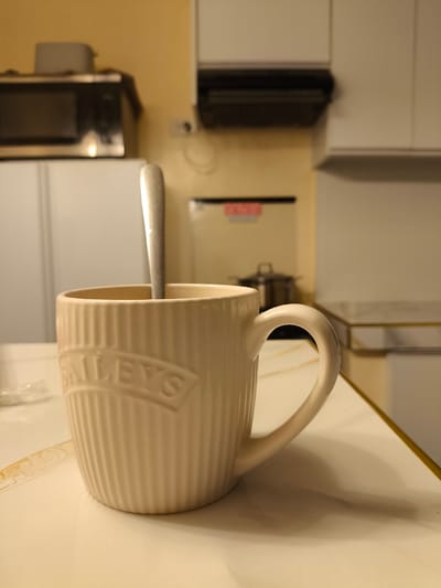The image shows a close-up of a beige mug with vertical ribbing and the word "BAILEY'S" embossed on its side. A silver spoon is placed inside the mug, partially submerged. The mug is sitting on a light-colored surface, possibly a countertop, which has a subtle marbled pattern.

In the background, a kitchen setting is visible. To the left, a microwave is perched on top of a refrigerator. Further back, a kitchen hood is mounted above a cooking area. The walls are a warm yellow hue, and there are kitchen cabinets. The overall lighting suggests it might be indoors, possibly in the evening or on an overcast day, given the warm artificial light.

There are no people visible in the image. The activity appears to be preparation or consumption of a beverage, indicated by the mug and spoon. The presence of kitchen appliances and cabinetry confirms the setting as a kitchen, likely in a home. No specific time of day or weather conditions are discernible beyond the interior lighting. No emotions are conveyed as the image focuses on inanimate objects. The text "BAILEY'S" is clearly visible on the mug. The location context provided is Imus, Philippines.