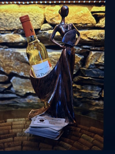 The image displays a close-up of a wine bottle held by a bronze-colored sculpture of a person with an outstretched arm. The sculpture appears to be abstract, with flowing lines and a stylized form. The wine bottle is made of clear glass and has a red foil capsule on the neck and a white label. Visible text on the label reads "LA PASSITO" and "LUCIEN ARKAS CILARI."
The background consists of a textured surface that resembles rough-hewn stone or rock formations in varying shades of brown and gray. To the right of the sculpture and bottle, a wall or surface is covered with numerous wine corks, some of which have visible markings or logos. The lighting is dramatic, with strong highlights and shadows that accentuate the textures of the background and the sculpture. The overall impression is one of an artistic display or still life.
The orientation of the image is landscape, with the subjects positioned diagonally. The quality of the image suggests it may have been captured from a screen or a photograph with a slightly grainy texture. No specific time of day, weather conditions, or emotions are discernible. The location context of Eyüpsultan, Turkey, does not provide immediate visual cues within the image itself.