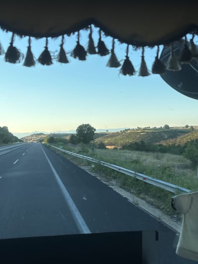 The image shows a view from inside a vehicle looking out onto a highway in Avra, Greece. The highway stretches into the distance, with rolling hills and sparse vegetation on either side. The sky is clear and blue. A metal guardrail runs along the right side of the highway. The top of the image shows a fringe of decorative tassels hanging from the vehicle's interior. There are no people visible in the image.