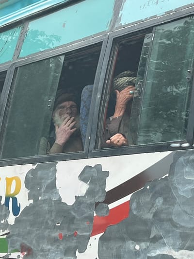 The image shows a close-up view of the windows of a bus. Through one window, an elderly man with a white beard is visible. He is wearing a turban and appears to be smoking, with his hand raised to his mouth. Through another window, a second person, also wearing a turban, is seen with their hand covering part of their face. A turquoise ring is visible on the ring finger of their right hand. The bus exterior is worn, with peeling paint and visible rust. Reflections of trees are visible in the bus windows. The image is taken from a slightly low angle, looking up at the bus. The context of the city of Kullan, India, is not directly depicted in the image itself, but the provided metadata indicates this location.