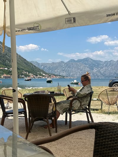 In Škaljari, Montenegro, a woman sits at a table under an umbrella, looking at her phone. She is wearing glasses and a patterned shirt. In front of her are drinks and a glass ashtray. The table is made of wicker and metal. Behind her, more wicker chairs are visible. The scene is outdoors, with a body of water and mountains in the background. Boats are scattered on the water. The umbrella has logos on it, including "TUBORG OPEN TO W". The sky is blue with some clouds.