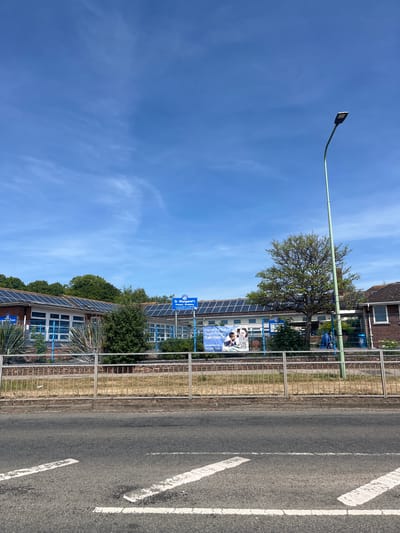 The image depicts St. Margaret's Primary School in East Suffolk, United Kingdom, as indicated by the signage. The school building is partially visible, featuring solar panels on the roof. A metal fence separates the school grounds from the adjacent road. The road has white painted markings. A lamppost stands to the left. The sky is predominantly blue with sparse clouds. A tree is visible behind the school building. A banner is attached to the fence, advertising teacher training.