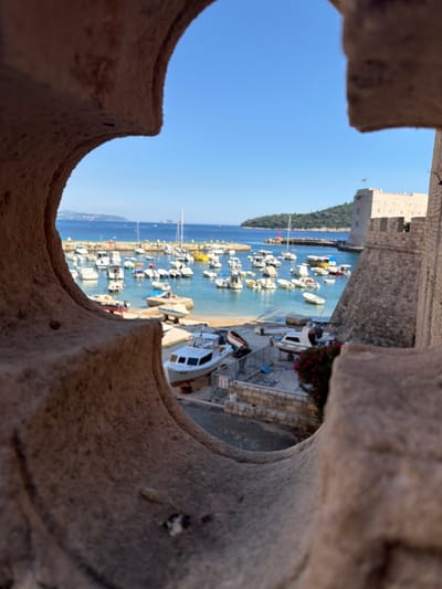 This is a photograph taken through a stone aperture, looking out at the harbor of Dubrovnik, Croatia, near Fort Lovrijenac. The aperture is in the foreground, with rough, textured stone on the left and right edges of the frame, partially obscuring the view of the harbor.
The harbor itself is filled with numerous small boats and yachts, moored in clear, turquoise water. The boats are of various sizes and colors, with many being white. Some boats are docked, while others are floating. The background of the image features a lush green hillside with more buildings visible in the distance, and the bright blue sky overhead. To the right of the harbor, a section of the stone fortifications of Dubrovnik is visible, with crenellated battlements and stone walls. A single flag flies from the top of the fortification. The overall scene depicts a sunny day in a picturesque coastal city.