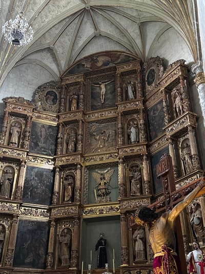 The image shows the ornate retablo of the San Miguel Arcángel church in Huejotzingo, Mexico. The retablo is a large, gilded altarpiece featuring numerous sculptures and paintings depicting biblical scenes and figures. In the foreground, a large crucifix with a statue of Jesus Christ is prominently displayed, with a sign bearing the inscription "INRI" attached to the cross. Behind the crucifix, the retablo rises in multiple tiers, showcasing a complex arrangement of niches containing statues of saints and angels, as well as painted panels. The architectural style of the church's ceiling, with its ribbed vaults and decorative elements, is visible in the upper portion of the image. The overall scene evokes a sense of religious devotion and historical artistry.