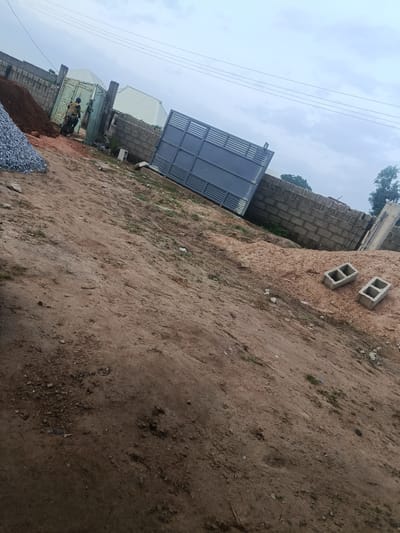 An exterior, eye-level, medium shot shows a construction site on a cloudy day. In the foreground, a pile of red dirt is on the right, with two concrete blocks and a pile of grey gravel on the left. In the midground, a person on a motorcycle drives past a light green gate and a grey metal gate, past a cinder block wall and two white-roofed buildings. In the background, telephone wires are visible against the sky.