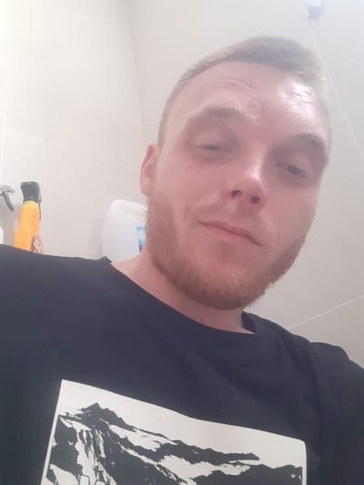 A close-up, slightly angled, selfie-style photograph depicts a light-skinned man with short, light brown hair, a light brown beard, and blue eyes. He is wearing a black t-shirt with a white graphic of a mountain range printed on the front. The man's head and upper torso are visible. The background is a plain, light-colored tiled wall. To the left of the man, partially visible, are two bottles of cleaning products. The overall lighting is bright and diffused, suggesting an indoor setting, possibly a bathroom or utility room. The man's facial expression is neutral, and he is looking directly at the camera. There is no discernible information in the image to determine the specific location within Tczew, Poland.