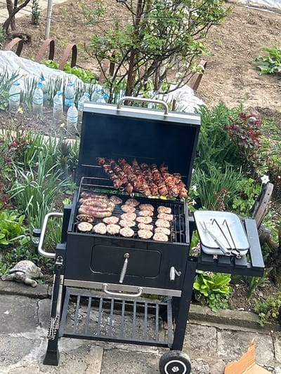 The image shows a black charcoal grill outdoors, likely in a yard. The grill is loaded with various meats: skewers on the left and rows of what appear to be sausages and burger patties on the right. The setting appears to be residential, with plants and stone paving visible in the background. There are water bottles and gardening tools. The location is in the city of Bukovlak, Bulgaria. There are no people visible in the frame.