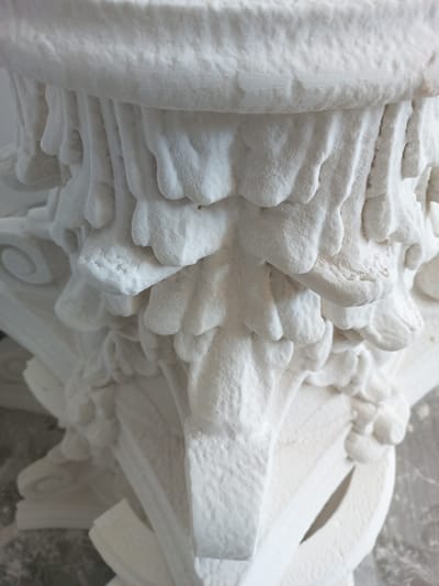 The image shows a close-up, detailed view of a white, intricate sculpture, possibly made of plaster or a similar material. The sculpture features ornate, layered elements that resemble foliage or drapery, with a rough, textured surface. The lighting suggests an indoor setting, possibly a workshop or studio, with soft, diffused light that highlights the texture and depth of the carving. The focus is entirely on the sculptural details, with no people or discernible activity. The background is blurred and muted, directing all attention to the subject. The composition emphasizes the craftsmanship and intricate design of the piece, with shadows and highlights defining the sculptural forms. There is no visible text in the image. The location context provided is Durlești, Moldova, which might suggest the origin or creation place of this artwork.