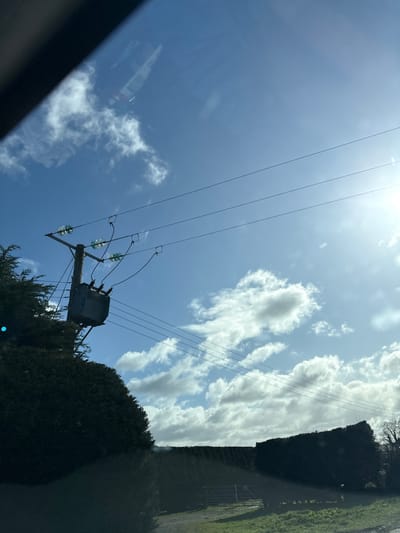 The image presents a daytime outdoor scene in Ruyton-XI-Towns, United Kingdom, captured from inside a vehicle, indicated by reflections in the upper left. A wooden utility pole stands prominently on the left, supporting a grey, cylindrical electrical transformer. Several green-tinted glass insulators are visible on the pole, connecting the transformer to three parallel overhead power lines stretching across the upper frame. Dark green, dense evergreen hedges and trees occupy the lower left and extend across the middle ground. In the lower right, a patch of green grass is visible, partially obscured by a lower hedge, behind which a metal gate and a dirt or gravel track are discernible. A bare deciduous tree is visible against the sky on the far right. The sky is clear blue with scattered white cumulus clouds, and the bright sun is present in the upper right, causing a lens flare effect. No individuals or active interactions are observed in the scene.